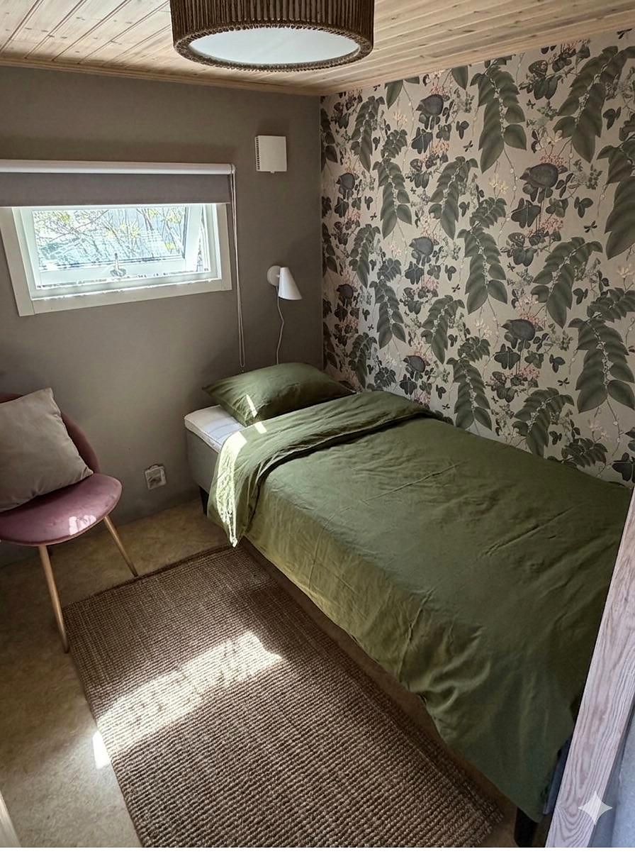 Einzelbett mit grüner Bettwäsche vor einer Wand mit Blumentapete, daneben ein rosa Sessel.