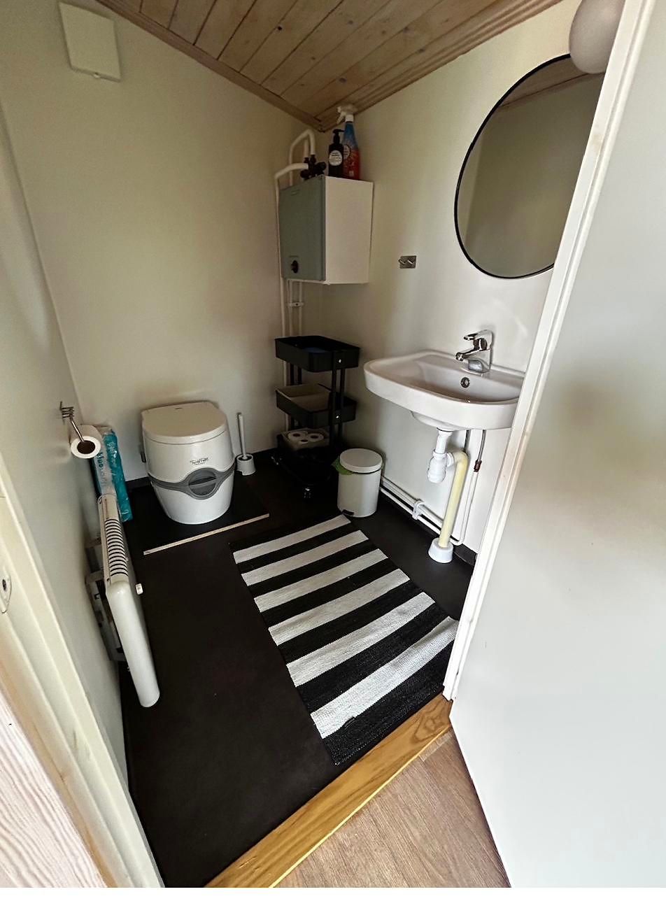Kleiner Raum mit Camping-Toilette, Waschbecken, Spiegel und gestreiftem Teppich auf schwarzem Boden.