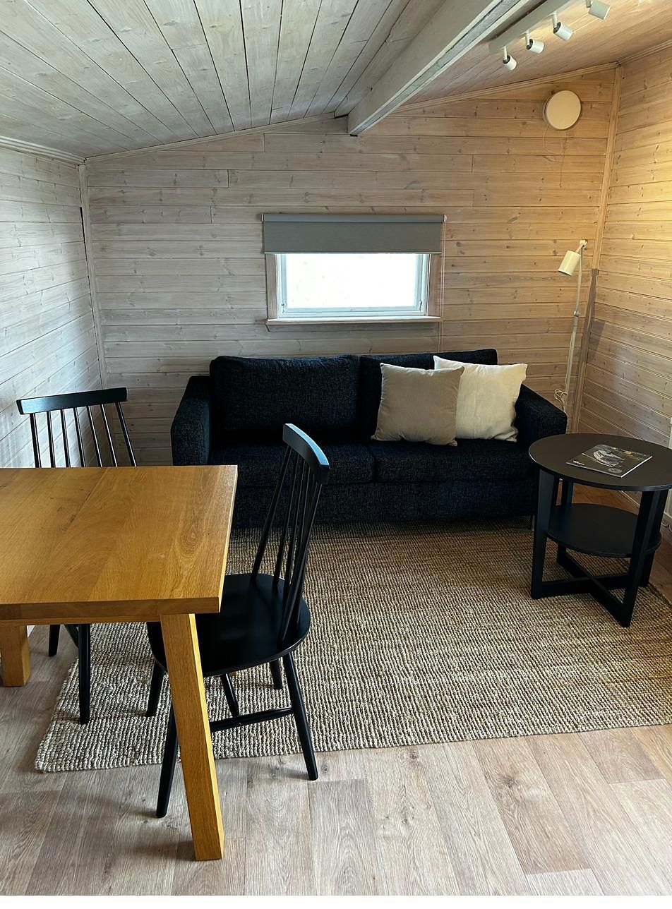Wohnbereich mit hellen Holzwänden, dunklem Sofa, Eichentisch und schwarzen Sprossenstühlen.